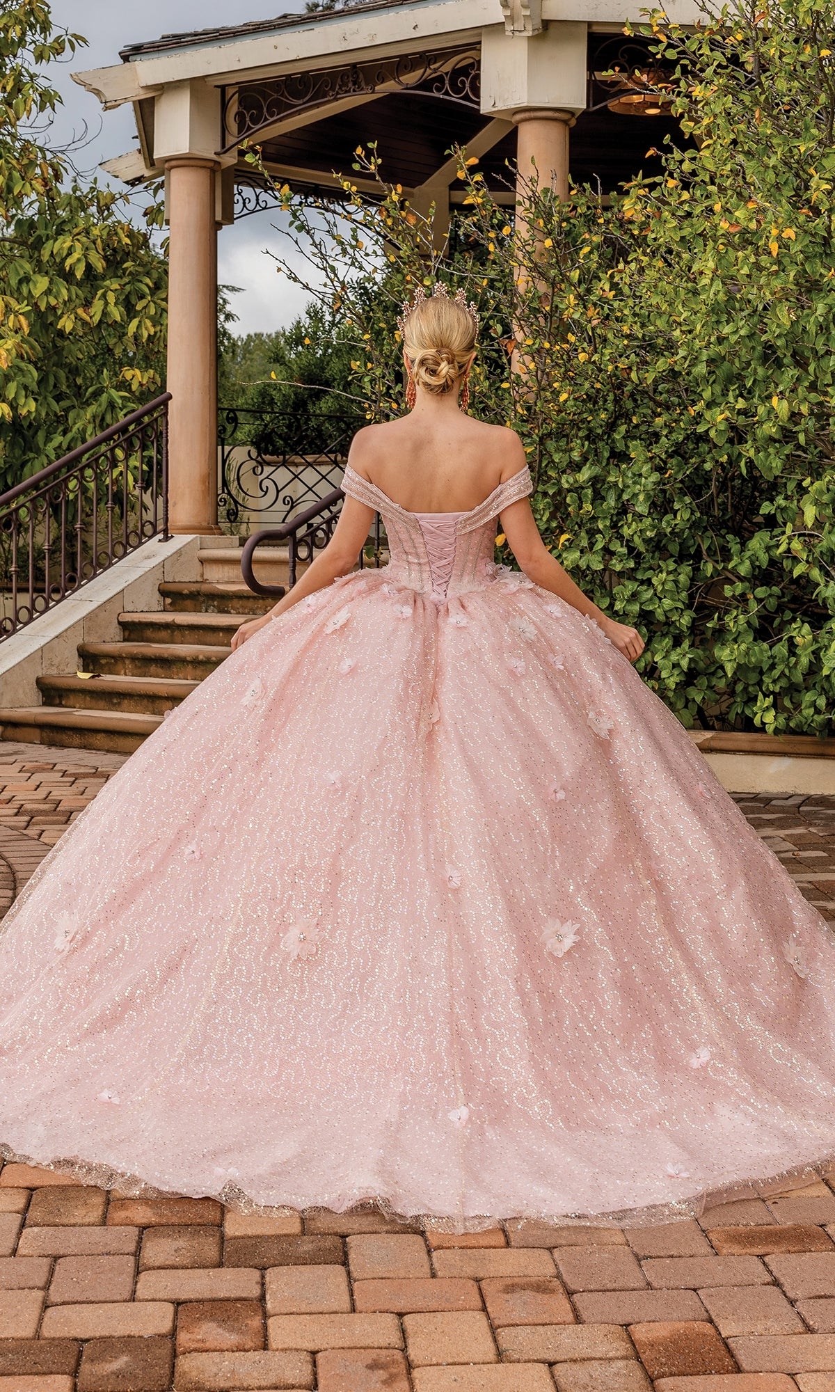 A woman stands outdoors in front of a gazebo, wearing the Quinceanera Dress 1868 By Dancing Queen—an off-the-shoulder pink gown—with her hair in a bun and greenery all around.